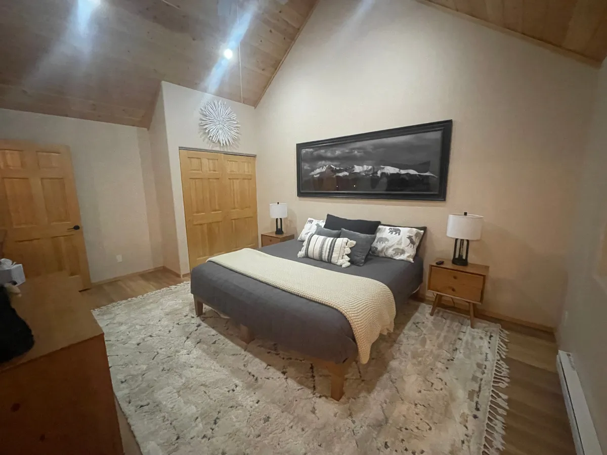A calming bedroom scene with a bed dressed in gray bedding, a striking black-and-white mountain photograph on the wall, wooden doors, and a decorative wall piece. The room is filled with soft, natural light.