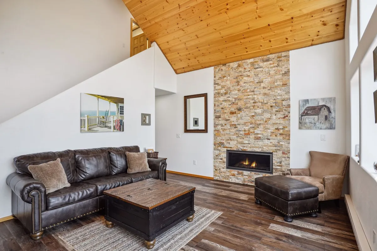 A spacious living room with a stone fireplace, leather sofa, wooden coffee table, and large windows that frame a stunning mountain view. The layout invites guests to relax, socialize, and enjoy both the cozy fire and the natural scenery outside.