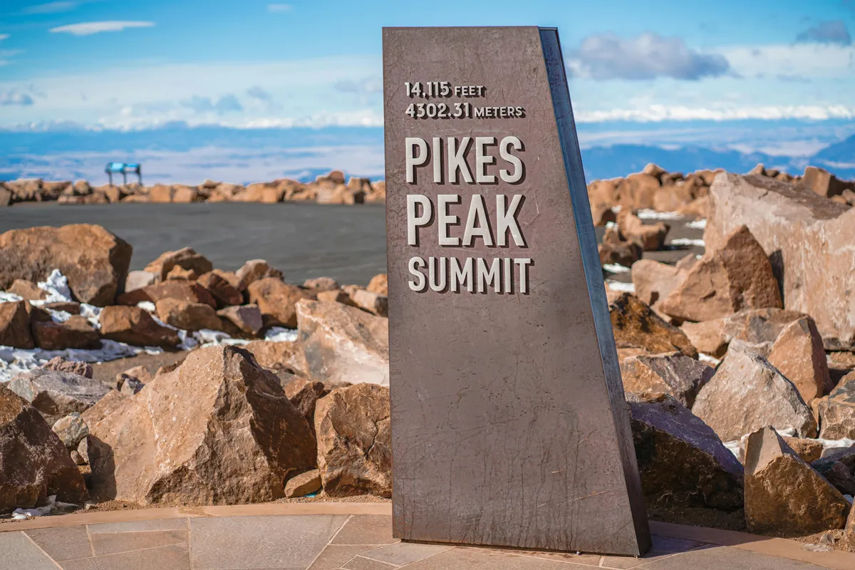 An inspiring exterior view at the Pikes Peak Summit sign, with rugged rocks and distant mountain vistas under open sky. This iconic scene evokes the thrill of exploration, hiking, and capturing sunrise or sunset photographs with awe-inspiring backdrops.