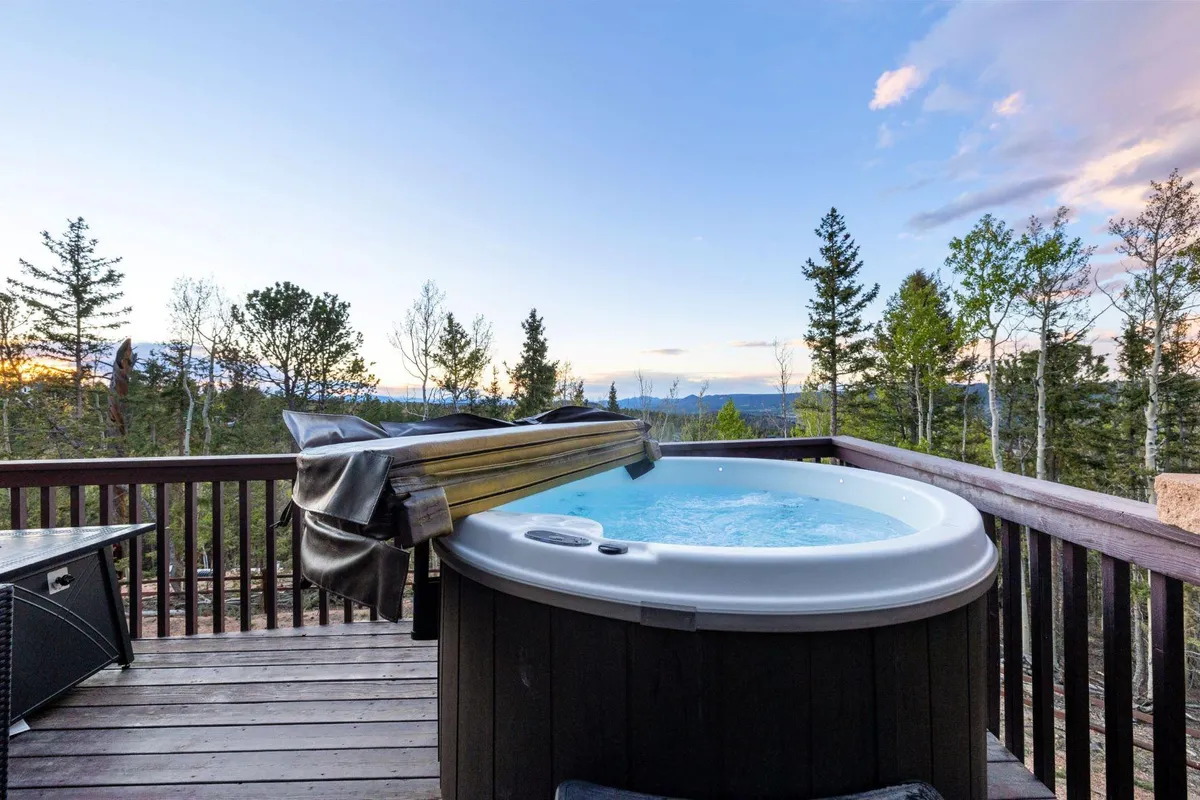 A tranquil scene presenting a hot tub positioned on a deck with surrounding trees and a vast mountain view in the distance. The ambiance invites guests to relax while immersed in natural beauty, ideal for both daytime and nighttime enjoyment.