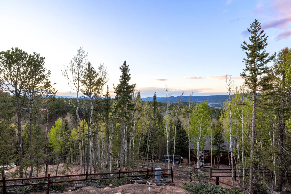 A panoramic view across the mountain landscape, featuring distant peaks, dense forests, and a wide, open sky. The fenced area in the foreground hints at proximity to the home while emphasizing opportunities for hiking, stargazing, and wildlife watching.