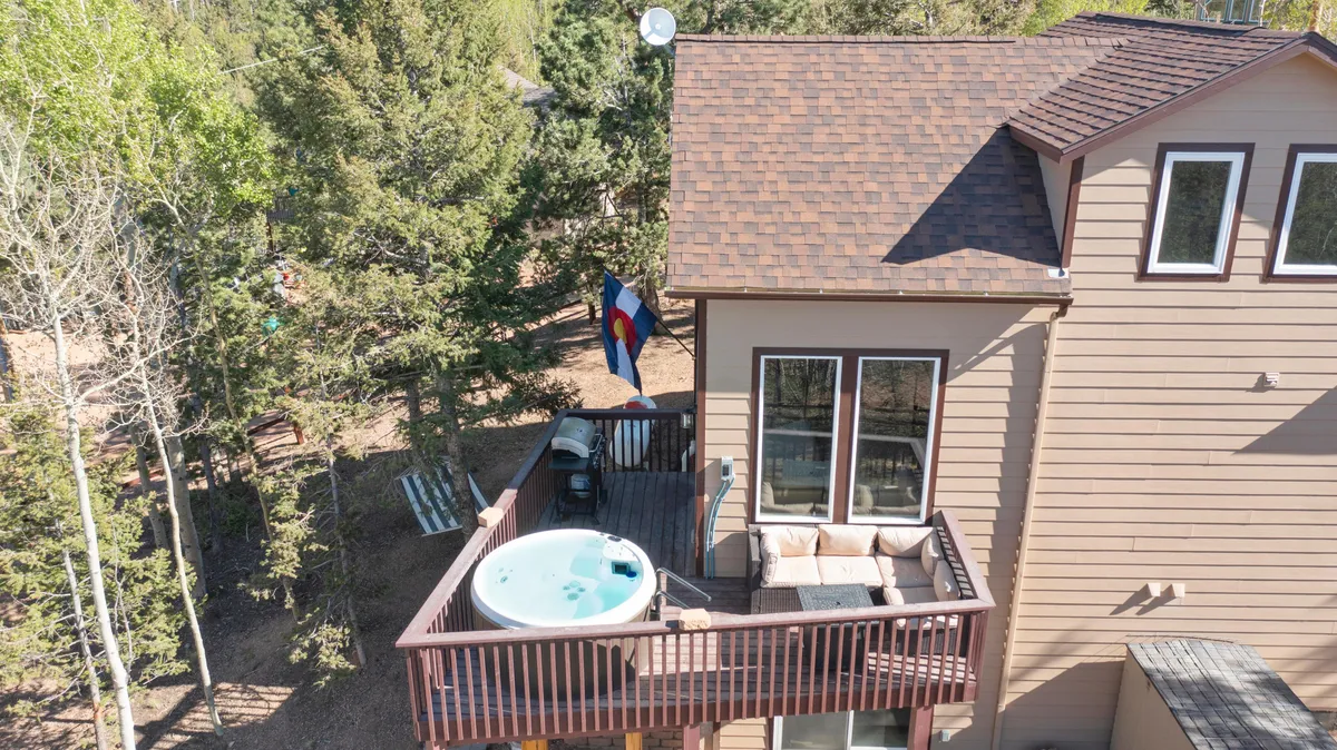 A serene exterior scene capturing a private hot tub nestled on a wooden deck, surrounded by trees and an outdoor seating area. The Colorado flag adds a regional touch, and the backdrop opens to tranquil natural views, suggesting a perfect spot for relaxation or stargazing.