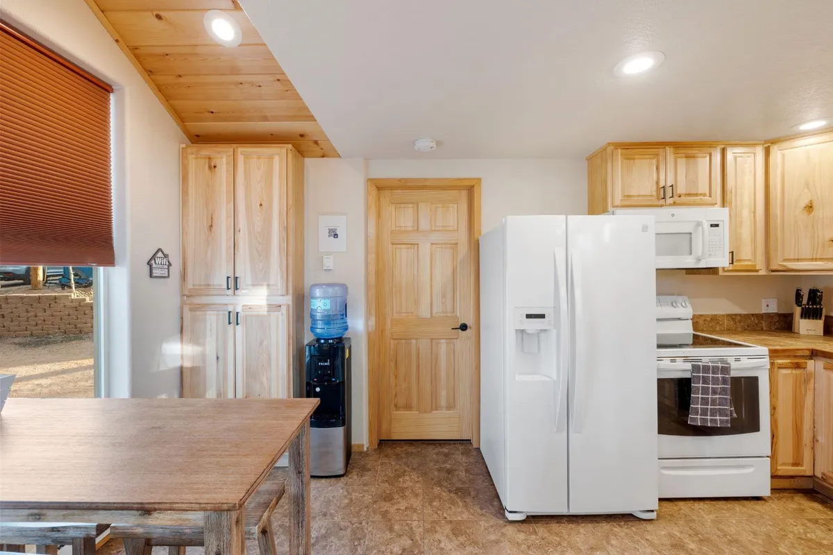 A bright kitchen and dining space featuring kitchen cabinets, a white refrigerator, a dining table set for meals, and a water cooler. The room is bathed in natural light, suggesting a cheerful environment for shared breakfasts or group dinners.