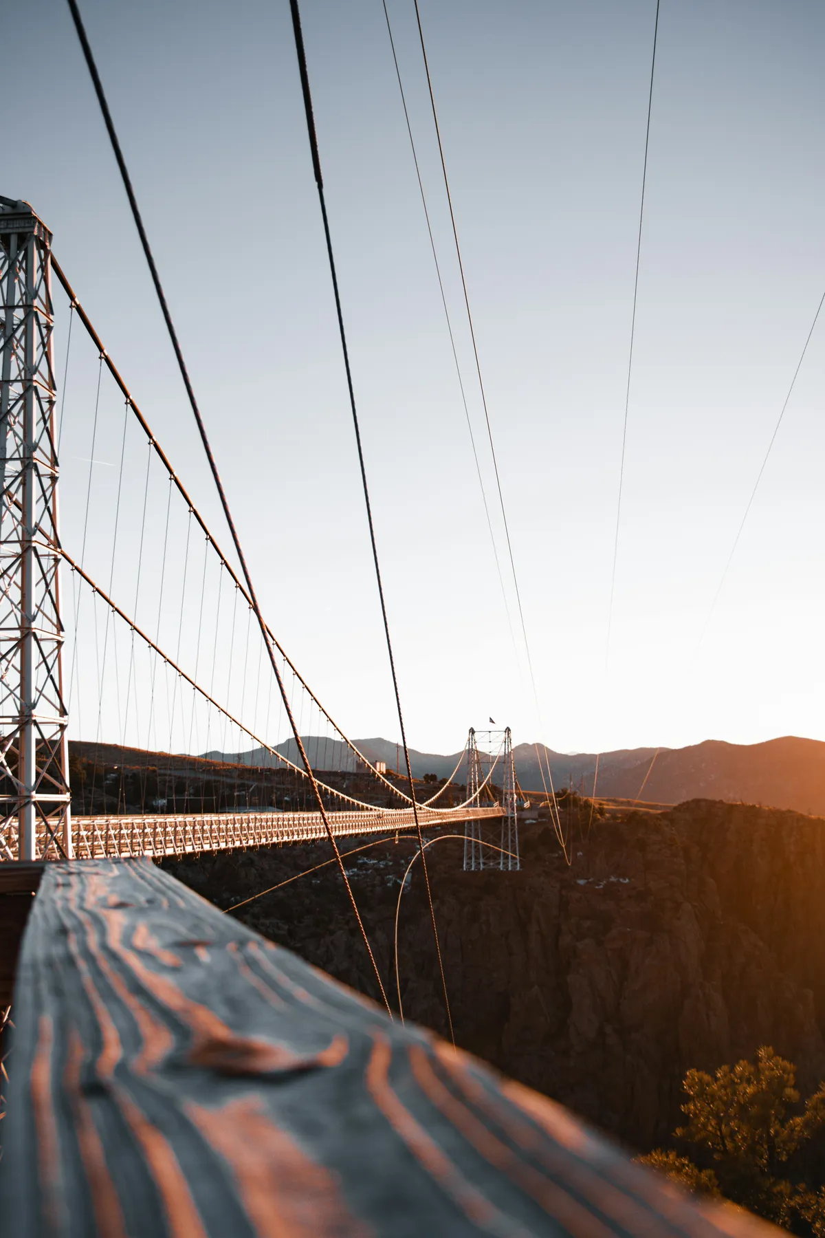 A suspension bridge with wooden railings stretches over a dramatic gorge, framed by a mountainous backdrop. The scene captures a moment of outdoor exhilaration and stunning views, ideal for adventure seekers.