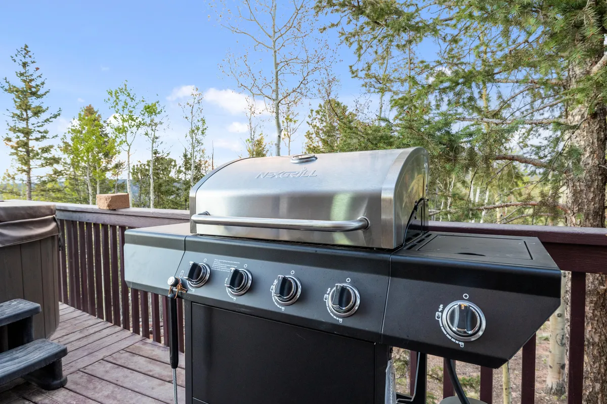 An inviting exterior image of a wooden deck with a modern grill, surrounded by lush trees. The setup is ready for outdoor dining and social gatherings, emphasizing the space for group activities and alfresco meals.