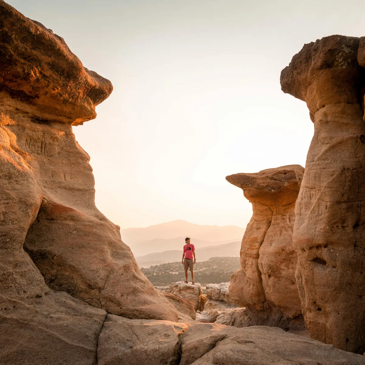 This image shows towering rock formations with a lone figure standing among the rocks, distant mountains on the horizon, and dramatic lighting that suggests late afternoon or sunset. The landscape evokes the rugged beauty and adventurous spirit of Colorado’s outdoors.