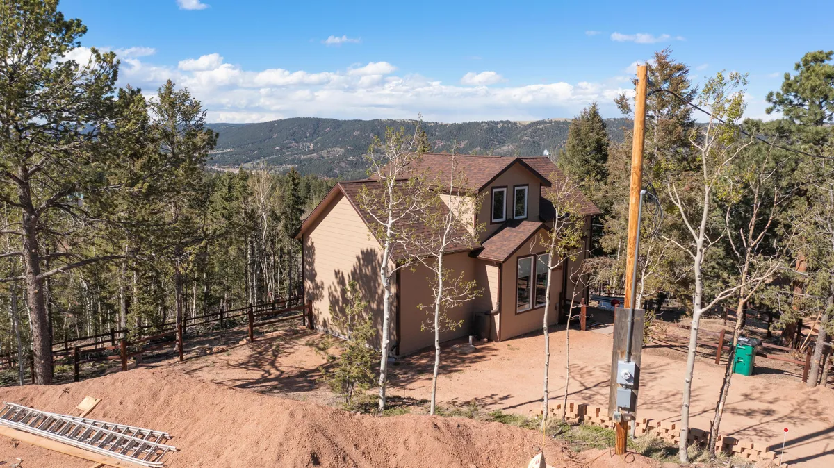 A wide exterior shot of the house nestled among tall trees with the dramatic mountain backdrop visible behind it. The image conveys the peaceful seclusion of the setting, with the home surrounded by nature and offering glimpses of the sky—perfect for stargazing at night or sunrise views.