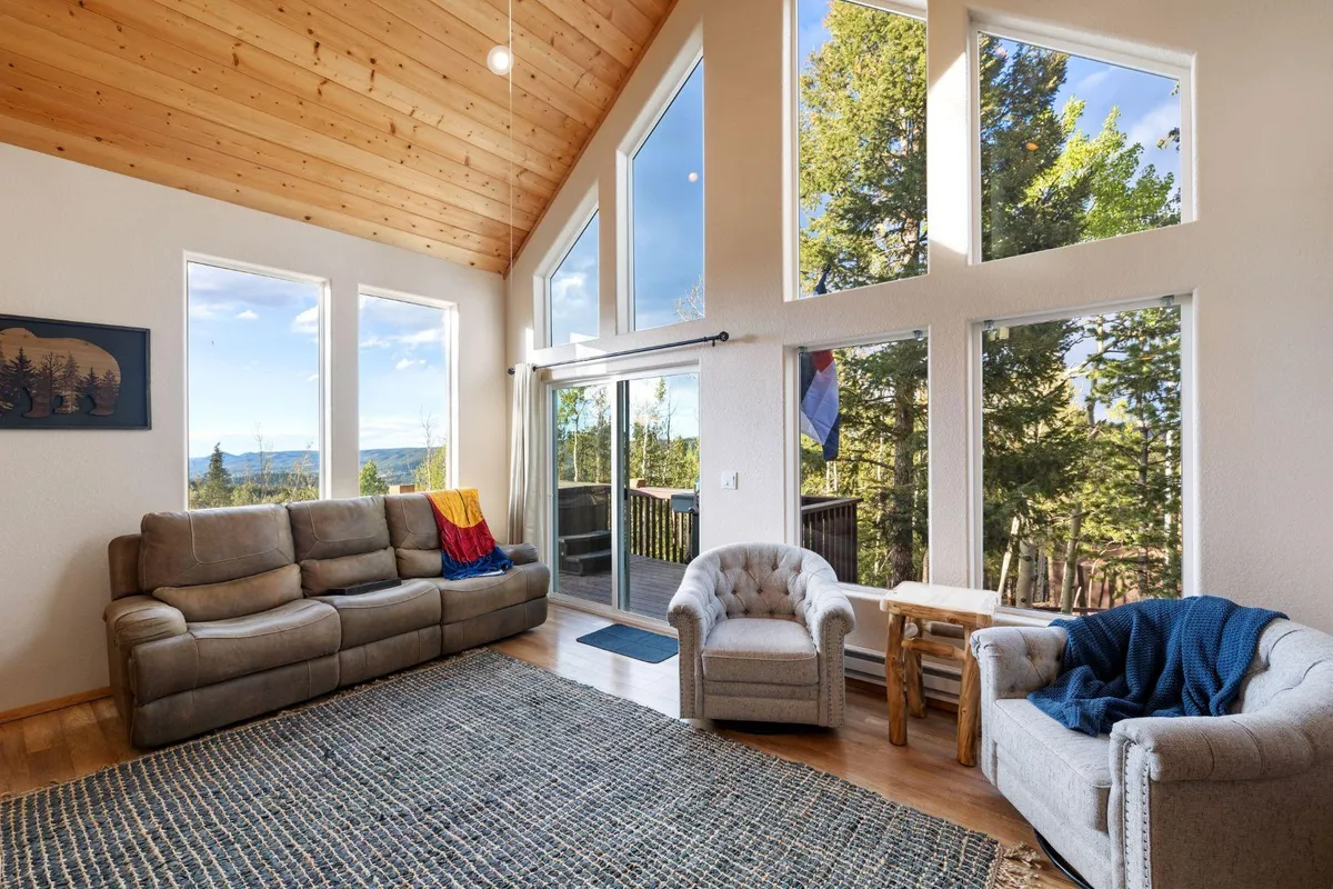 A welcoming living area featuring large windows that frame tree views, a comfortable sofa and chairs, a wooden coffee table, and a patterned area rug, all arranged to encourage group relaxation and conversation. The space feels open and inviting, perfect for communal gatherings and cozy evenings.
