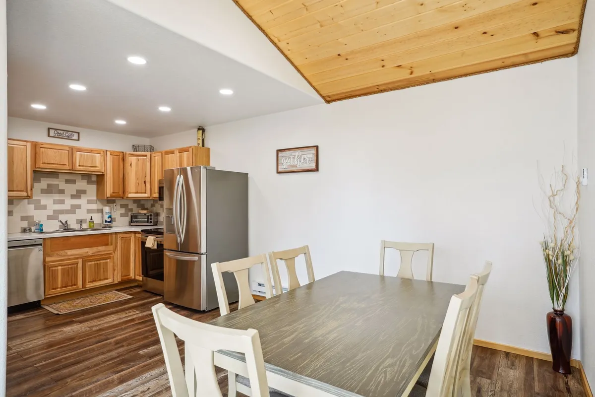 A welcoming dining area with a wooden dining table, kitchen cabinets, fridge, and decorative wall art. The space is designed for shared meals, cooking together, and entertaining, with plenty of room for groups.