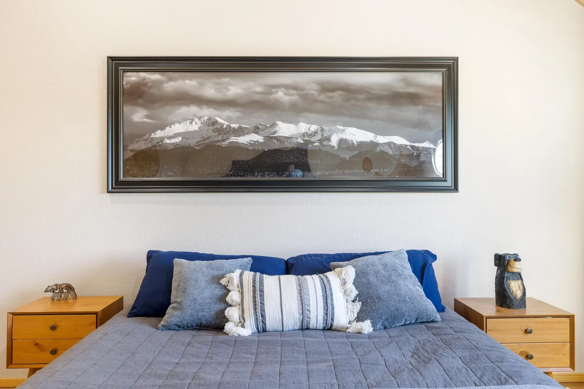 A serene bedroom featuring a bed with decorative pillows, mountain landscape artwork above the headboard, and nightstands adorned with sculptures. Natural morning light pours in, creating a peaceful and inviting atmosphere.