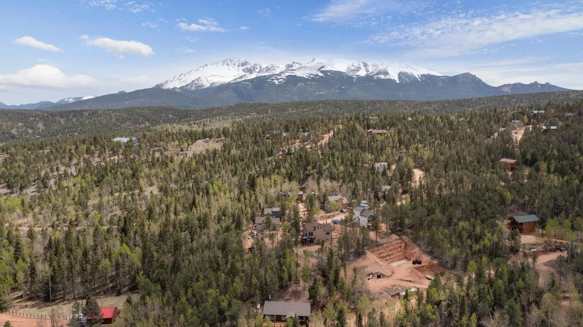 A sweeping view of snow-capped mountains rising above a dense forest with scattered cabins in the foreground. The scene radiates tranquility and offers a sense of untamed wilderness, ideal for wildlife observation and nature photography. The vast landscape invites viewers to imagine spotting deer or birds in their natural habitat.
